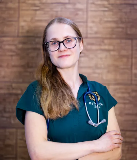Veterinarijos gydytoja Lina su akiniais ir stetoskopu prie medinės sienos